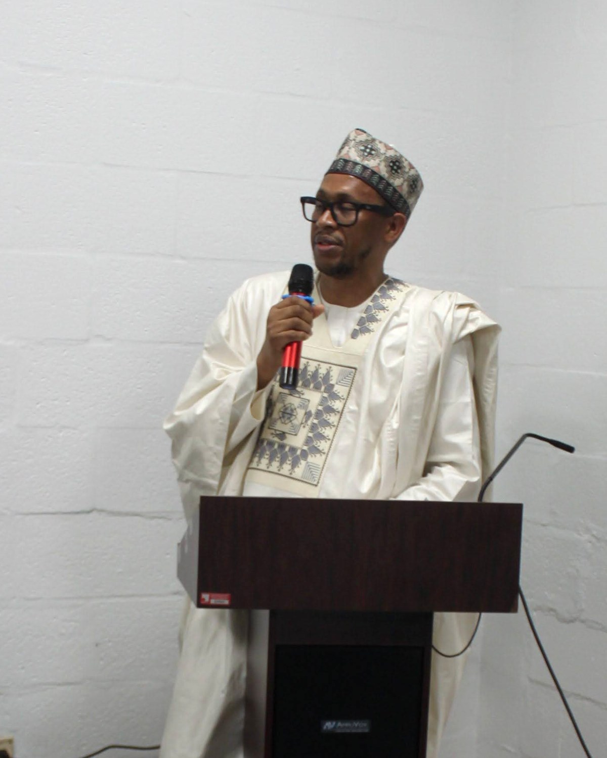 The president of FAMSO, in traditional white attire and embroidered cap, addressing the community from the podium